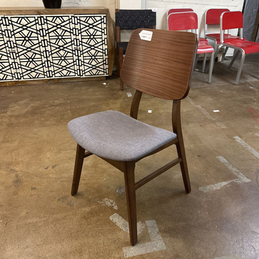 Wooden chair with gray cushioned seat on a concrete floor, with other chairs and a patterned wall in the background.