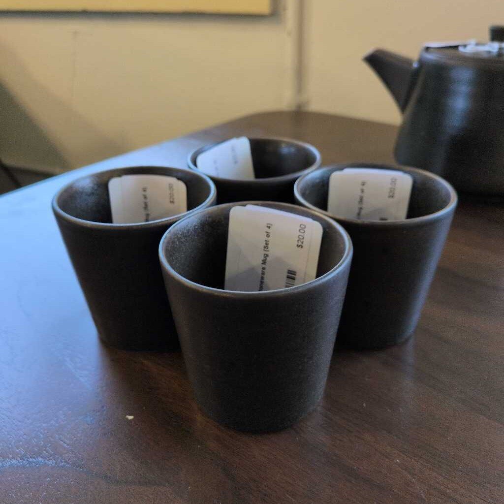 Four dark ceramic cups with labels on a wooden surface, accompanied by a teapot.