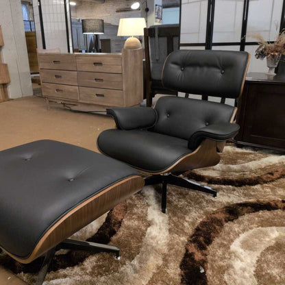 Black Eames-style lounge chair and ottoman on a patterned rug with furniture in the background.