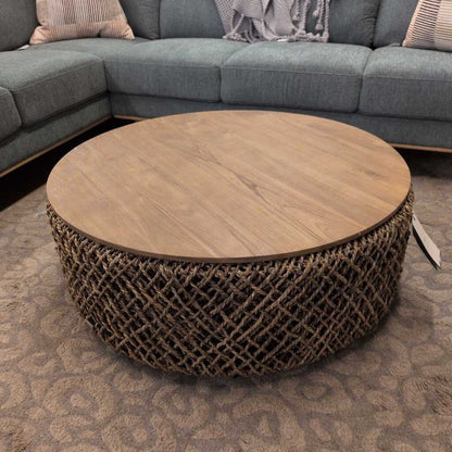 Round wooden coffee table with woven base in front of a gray sectional sofa.