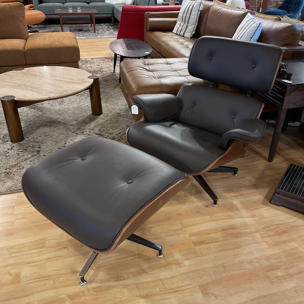 Black Eames-style lounge chair and ottoman on a wooden floor with furniture in the background.