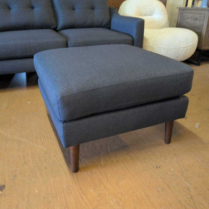 Gray upholstered ottoman on a wooden floor with a couch and chair in the background.