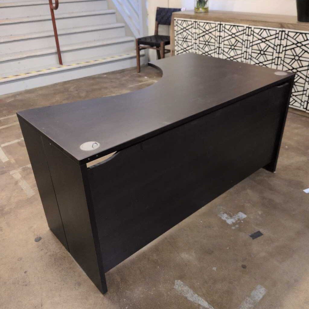 Black L-shaped desk in an indoor setting with a patterned wall and staircase in the background.