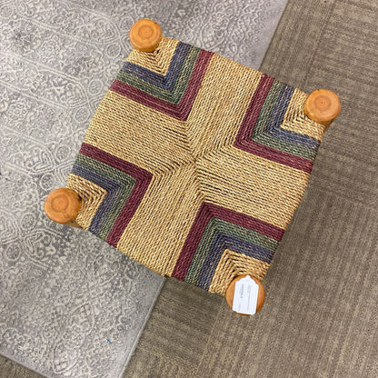 Small woven stool with a geometric pattern on a textured floor.