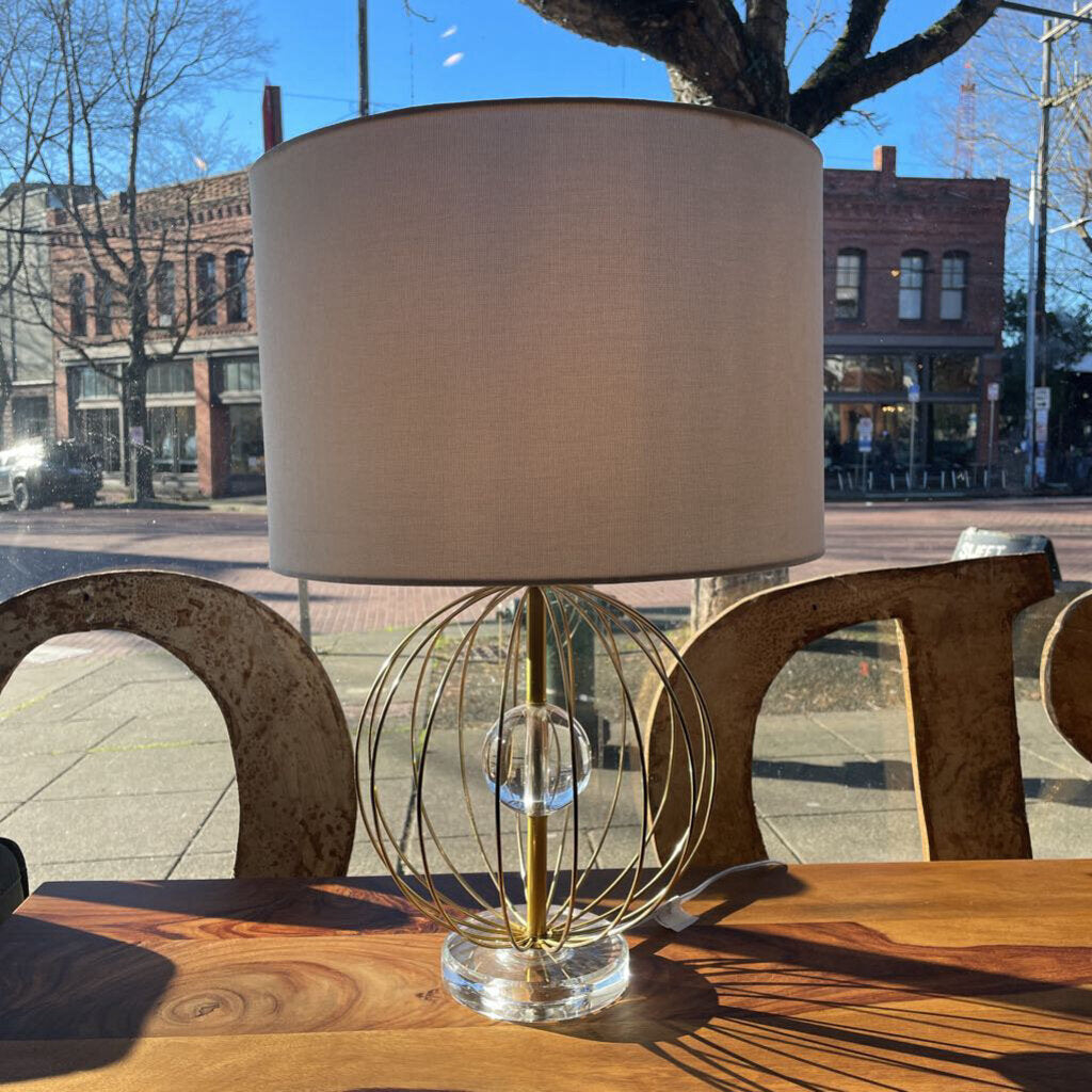 Table lamp with a clear glass base and beige shade on a wooden table outdoors.