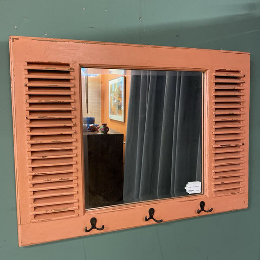 A rectangular wall mirror with a salmon pink distressed wood frame, featuring built-in louvers on the sides and three black metal hooks at the bottom