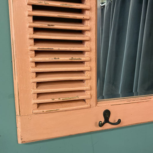 A rectangular wall mirror with a salmon pink distressed wood frame, featuring built-in louvers on the sides and three black metal hooks at the bottom