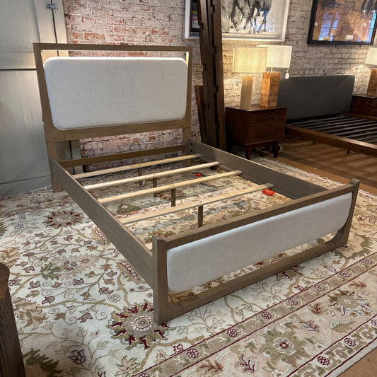 Wooden bed frame with white headboard on a patterned rug in a room with brick walls.