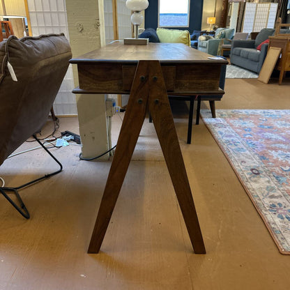 A slim, dark wood writing desk with a minimalist rectangular top, two open storage cubbies, and angled A-frame legs, shown on a neutral carpet.
