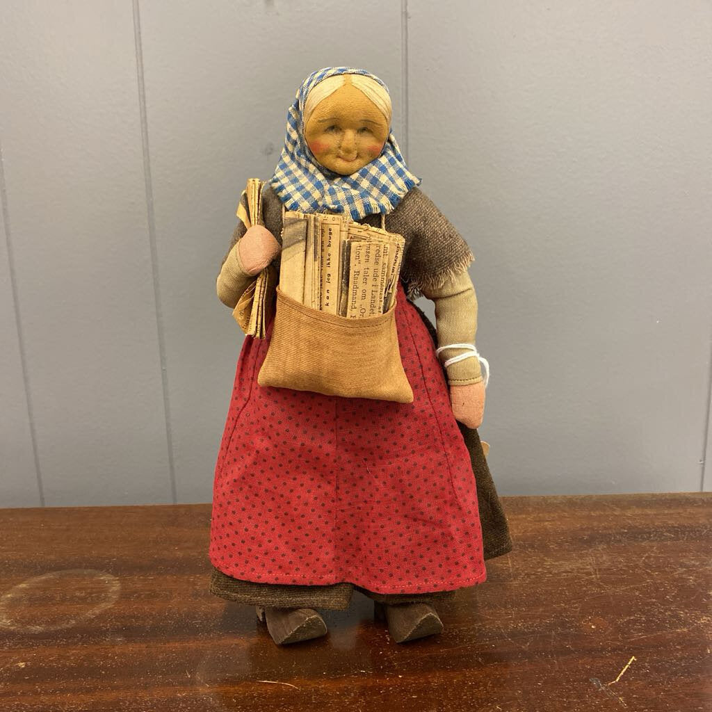 Vintage-style doll with a red dress, checkered headscarf, and newspaper bag on a wooden surface.