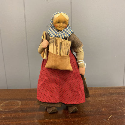 Vintage-style doll with a red dress, checkered headscarf, and newspaper bag on a wooden surface.