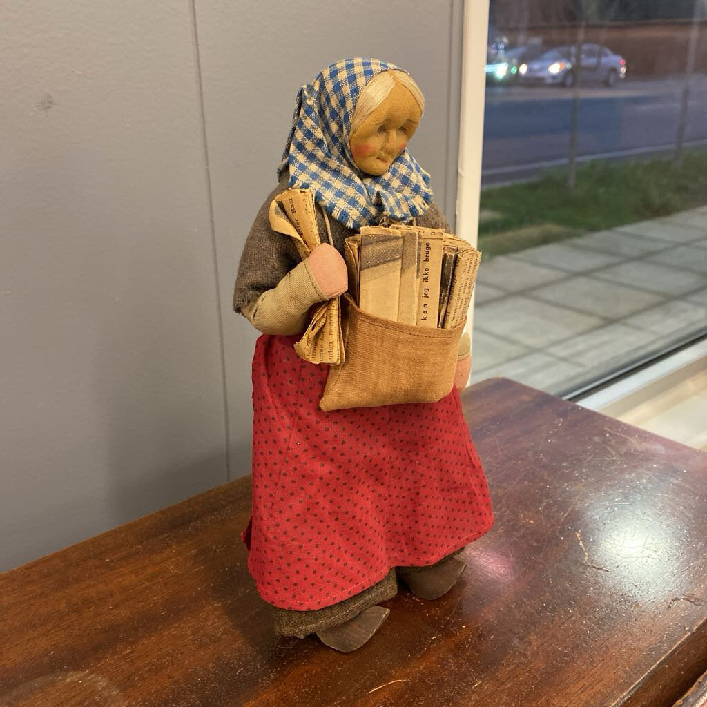 Vintage-style doll with a checkered headscarf and red apron holding a basket on a wooden surface.