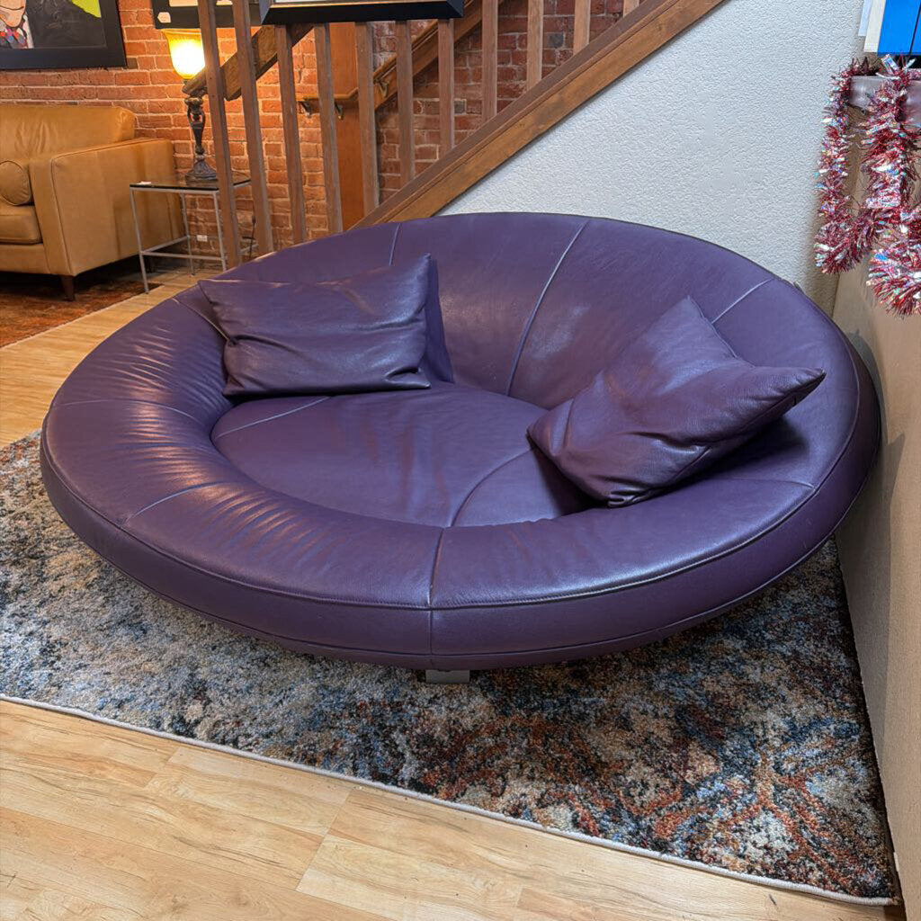 Round purple leather sofa in a living room setting with a staircase and lamp in the background.