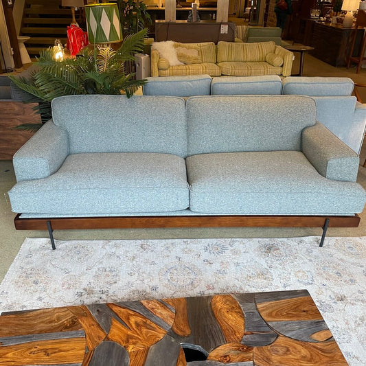  The piece is upholstered in a light seaglass or cool grey textured woven fabric, creating a soft, inviting aesthetic. The sofa features a low-profile frame and a deep two-cushion seat. Its structured silhouette is defined by wide, rectangular track arms and matching plush back cushions. 