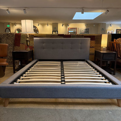 Gray upholstered bed frame with wooden legs in a showroom setting.
