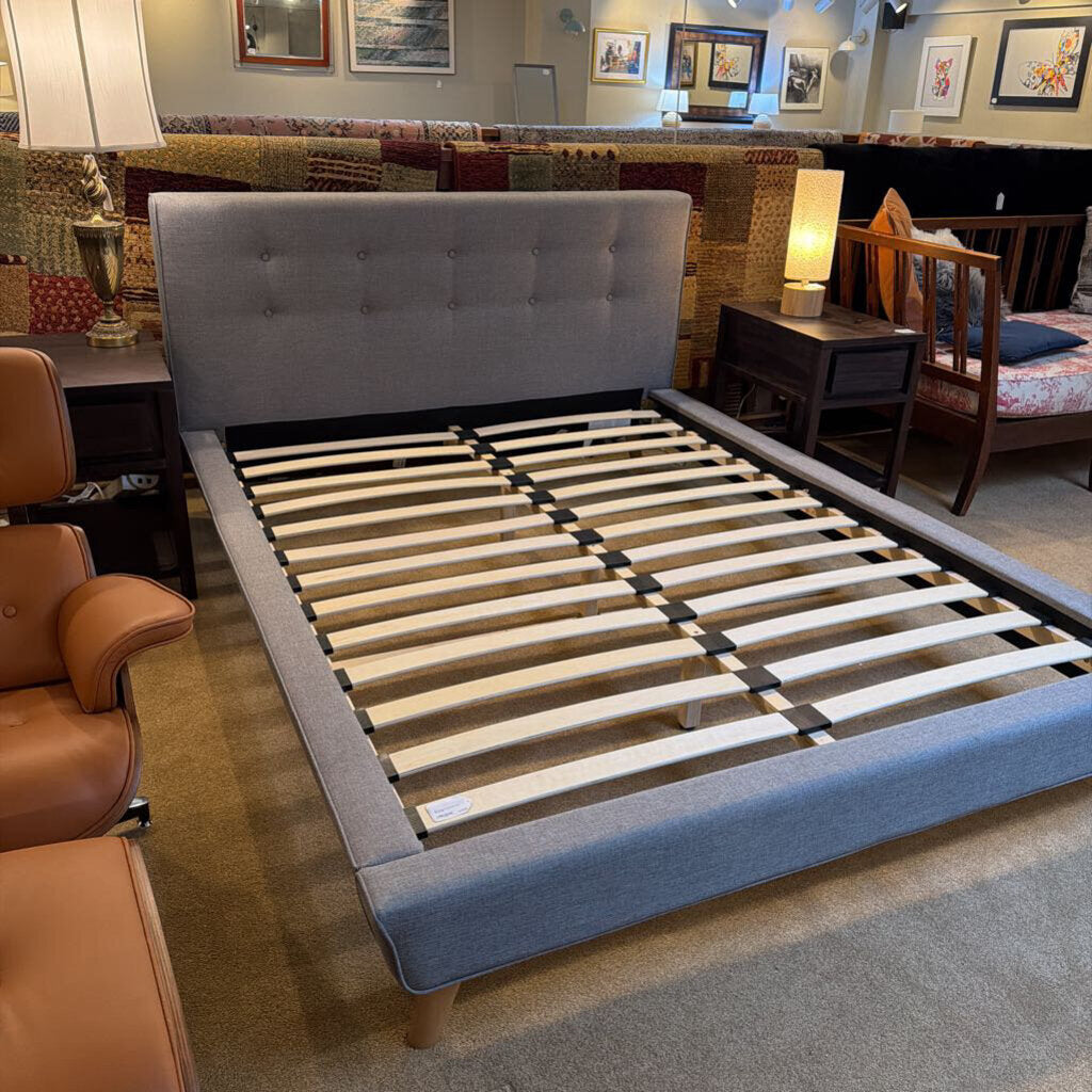 Gray upholstered bed frame with wooden slats in a showroom setting.