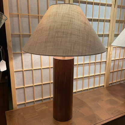 Table lamp with a brown cylindrical base and textured shade in front of a wooden panel.