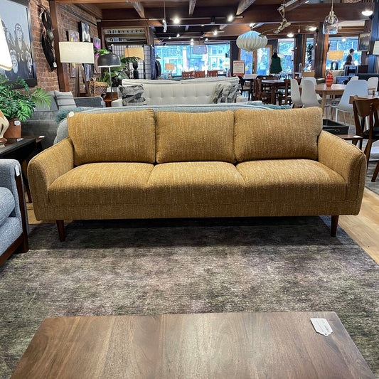  The piece is upholstered in a textured, woven fabric in a golden-wheat hue, characteristic of retro "mod" aesthetics. The sofa features slim, gently flared track arms and thick, plush back cushions, offering a balanced blend of comfort and structured style. 