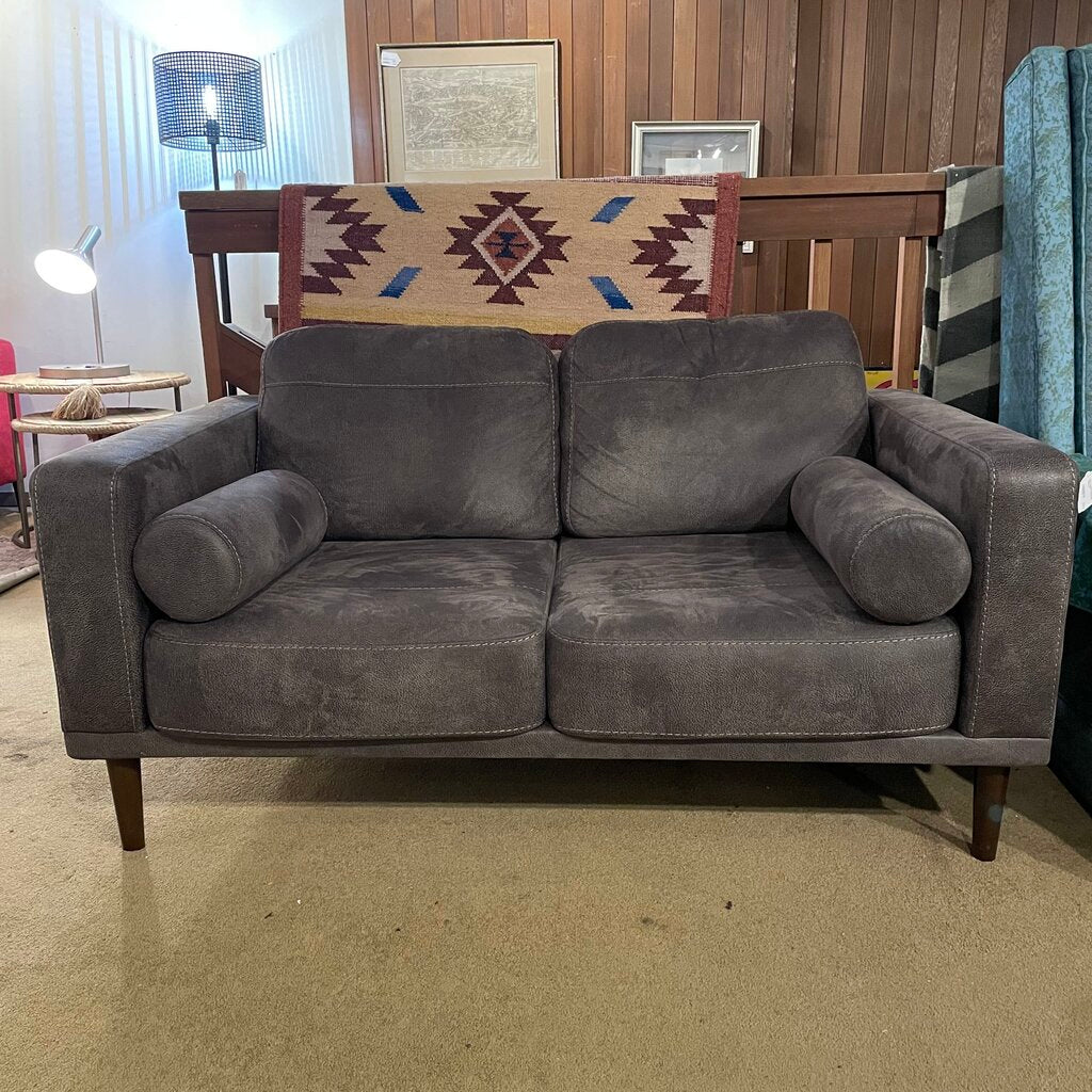 A dark grey, two-seater loveseat with a faux suede or velvet-like texture, featuring matching bolster pillows and tapered wooden legs, shown in a home decor setting