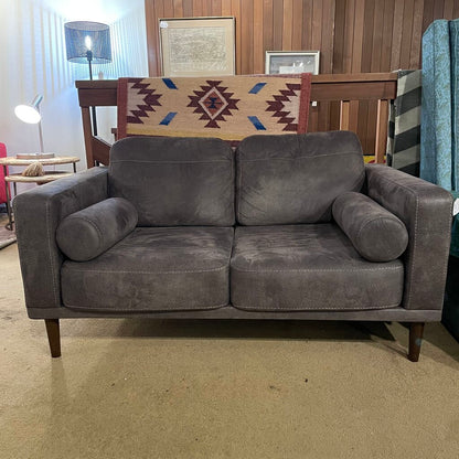 A dark grey, two-seater loveseat with a faux suede or velvet-like texture, featuring matching bolster pillows and tapered wooden legs, shown in a home decor setting