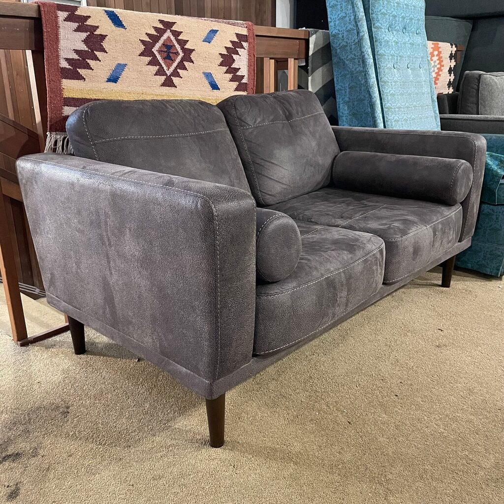 A dark grey, two-seater loveseat with a faux suede or velvet-like texture, featuring matching bolster pillows and tapered wooden legs, shown in a home decor setting