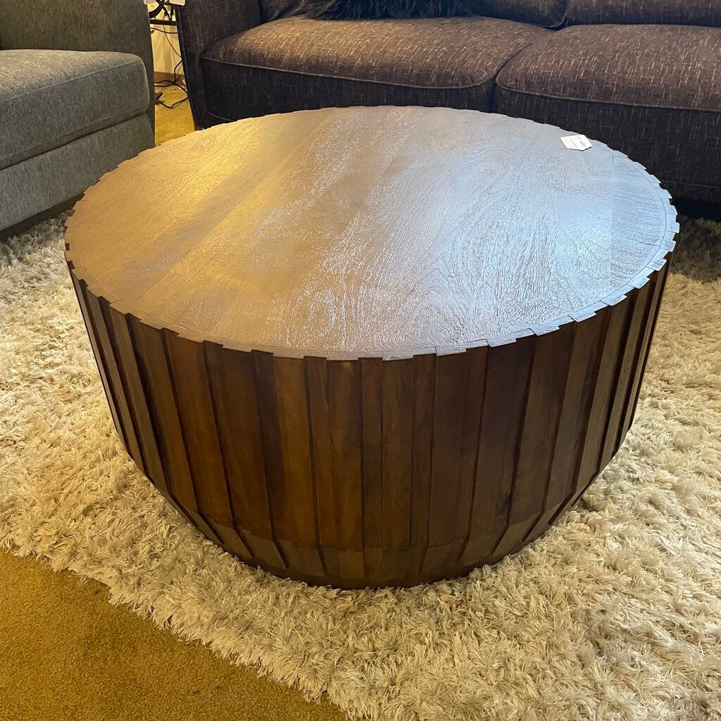 Wooden coffee table with a round top and ribbed cylindrical base in a living room setting.