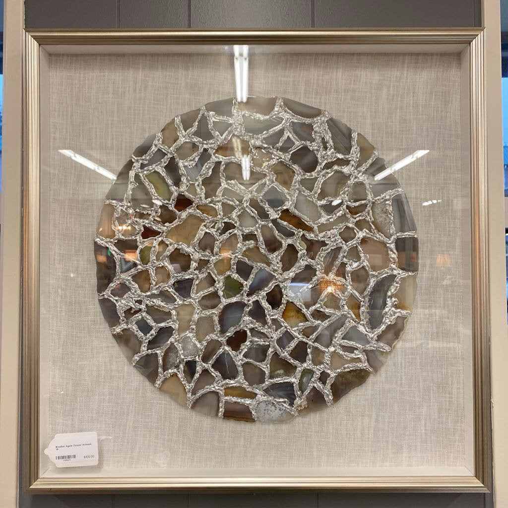 A framed wall art piece featuring a large circular arrangement of earth-toned agate slices connected by a textured silver-colored web, mounted on an ivory linen background