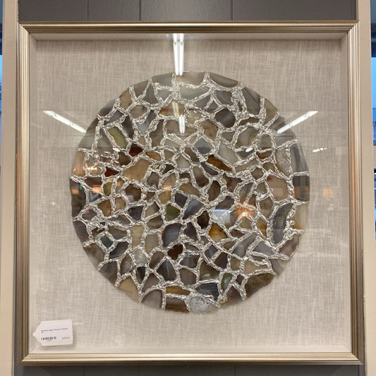 A framed wall art piece featuring a large circular arrangement of earth-toned agate slices connected by a textured silver-colored web, mounted on an ivory linen background