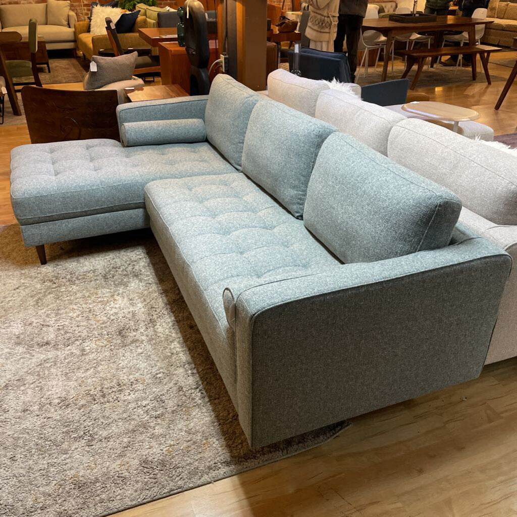 A light grey textured fabric sectional sofa featuring a left-arm-facing chaise with button-tufted seat cushions, two matching bolster pillows, and tapered wooden legs.