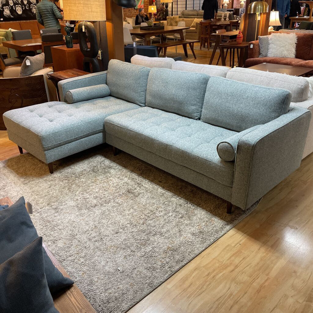 A light grey textured fabric sectional sofa featuring a left-arm-facing chaise with button-tufted seat cushions, two matching bolster pillows, and tapered wooden legs.