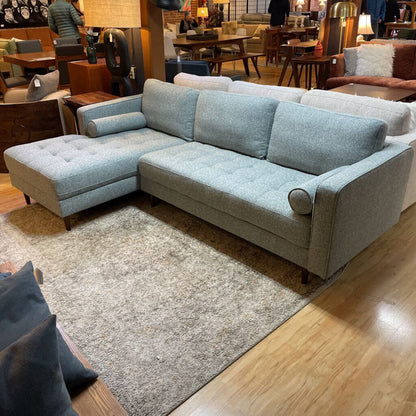 A light grey textured fabric sectional sofa featuring a left-arm-facing chaise with button-tufted seat cushions, two matching bolster pillows, and tapered wooden legs.