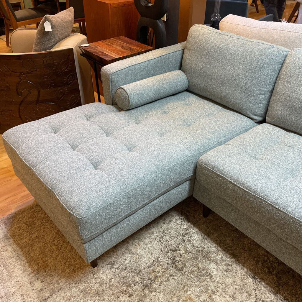A light grey textured fabric sectional sofa featuring a left-arm-facing chaise with button-tufted seat cushions, two matching bolster pillows, and tapered wooden legs.