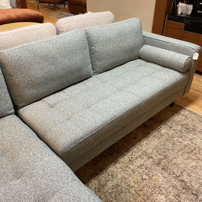 A light grey textured fabric sectional sofa featuring a left-arm-facing chaise with button-tufted seat cushions, two matching bolster pillows, and tapered wooden legs.
