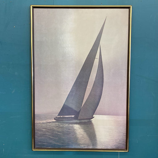 A vertical framed art piece depicting a large sailboat with towering grey sails cutting through calm water, shown under a misty sky with light reflecting off the sea's surface