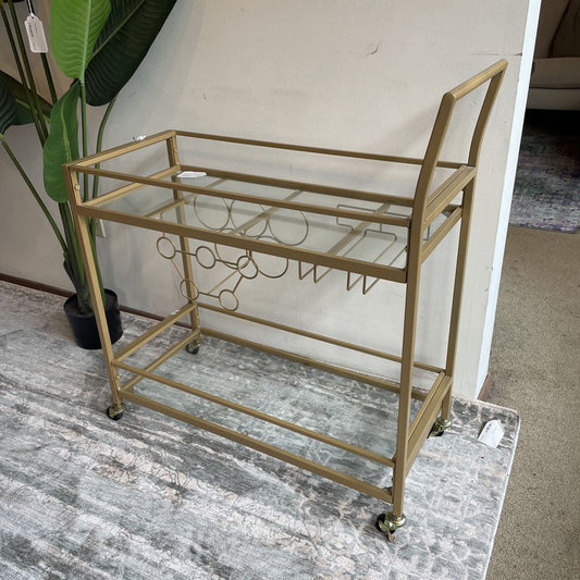 Gold bar cart with glass top and wine glass holder on a marble floor.