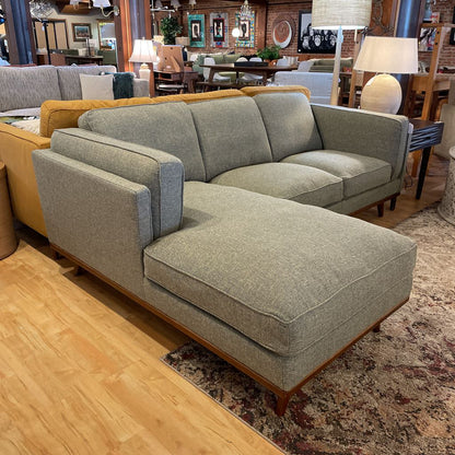 Gray sectional sofa in a showroom setting with wooden flooring and furniture in the background.