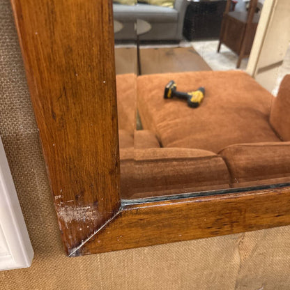 Wooden-framed mirror reflecting a brown couch and a drill on a carpeted floor.