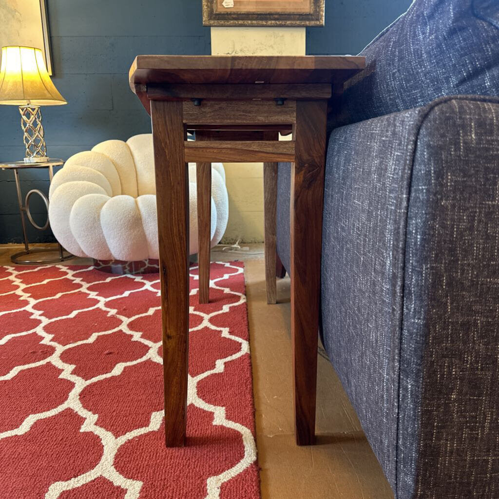 Wooden side table next to a gray sofa on a red patterned rug with a blue wall and white chair in the background.