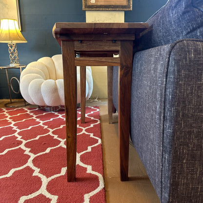 Wooden side table next to a gray sofa on a red patterned rug with a blue wall and white chair in the background.