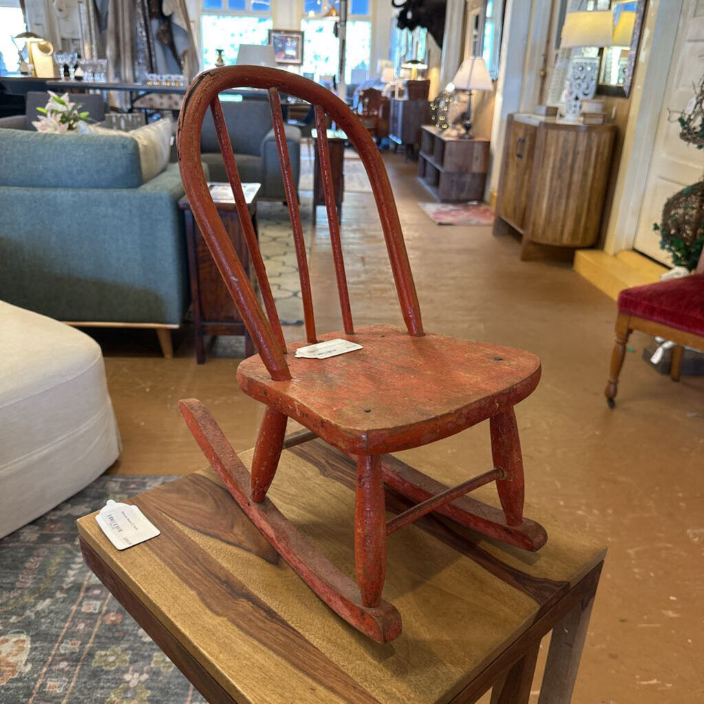 Red wooden rocking chair on a wooden platform in a store setting with furniture and decor.