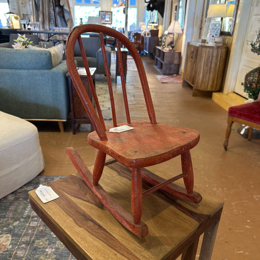Red wooden rocking chair on a wooden platform in a store setting with furniture and decor.