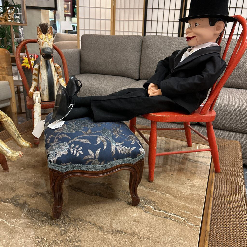 Mannequin in a suit sitting on a red chair with a blue cushioned stool in a room setting.