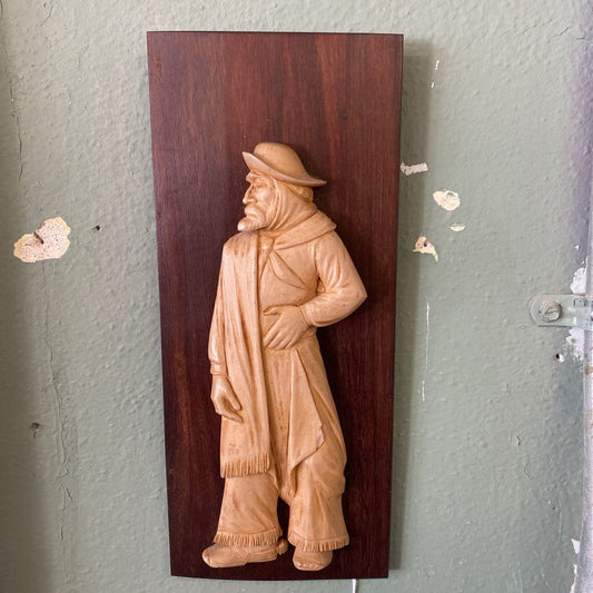 Wooden plaque with a carved figure of a person on a gray wall.
