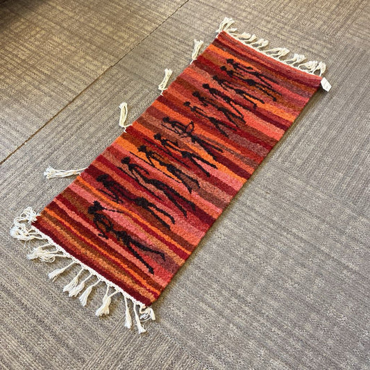 Small woven rug with red and orange stripes and white tassels on a gray floor.