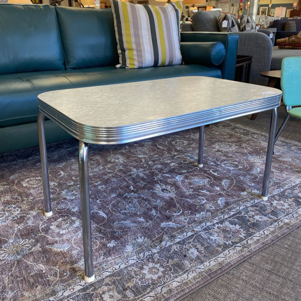 Metallic coffee table on a patterned rug with a teal couch and striped pillow in the background.