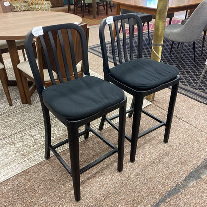 Two black bar stools with black cushions on a tiled floor.