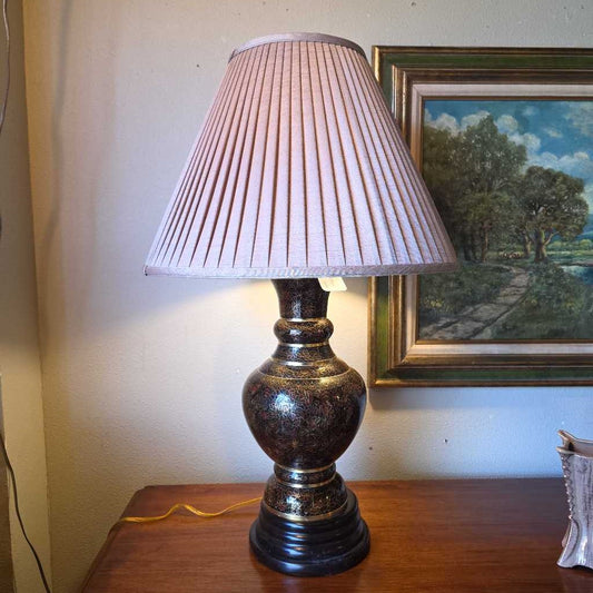 Table lamp with pleated shade on a wooden surface, with a painting in the background.