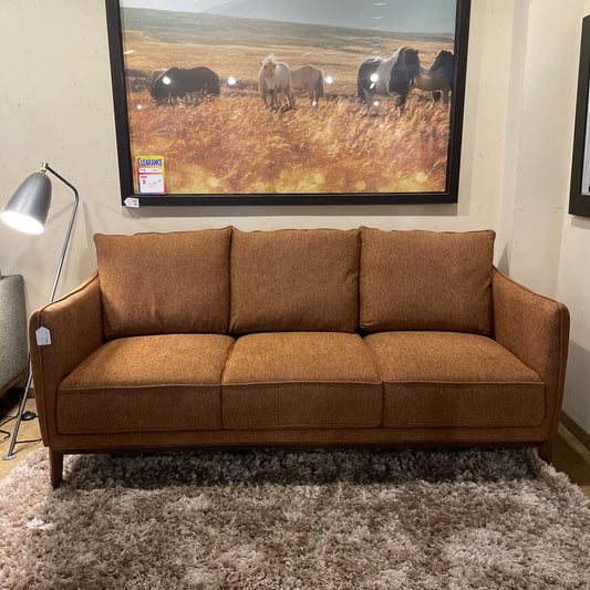 A burnt-orange textured three-seater sofa with three back cushions and a wooden base frame, resting on a high-pile shag rug