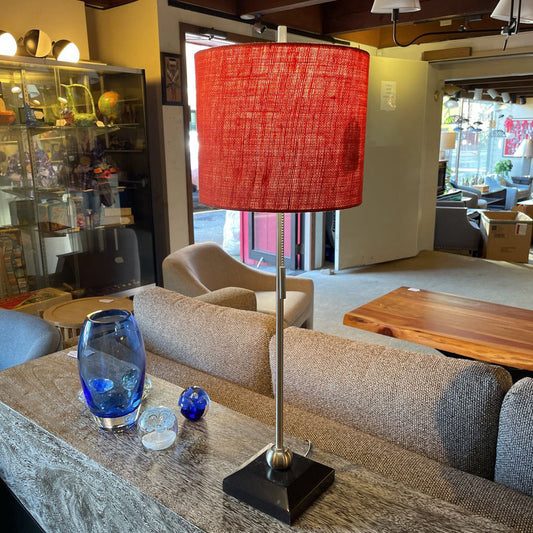 Red lampshade on a table in a living room setting with a couch and coffee table.