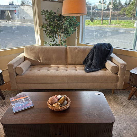 Beige sofa with a blanket and a wooden coffee table with a bowl and book in a room with large windows.